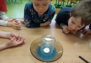 Dzieci obserwują eksperyment z niebieską wodą i świeczką.